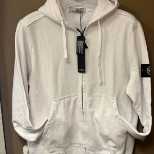 Vit hoodie från Stone Island - Säljer en stilren vit hoodie från Stone Island med dragkedja framtill och klassisk logopatch på vänster ärm. Hoodien har justerbar huva med snören och två stora fickor framtill. Tillverkad i mjukt bomullsmaterial för en skön känsla.