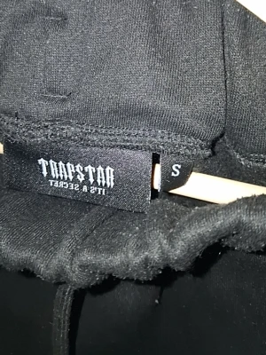 Svart Trapstar hoodie med tryck - Svart hoodie från Trapstar med huva och dragsko. Framsidan har ett stort Trapstar-tryck i rött, grått och vitt samt texten 'It's a secret' under. Klassisk känguruficka och ribbade muddar. Perfekt för en streetwear-look.