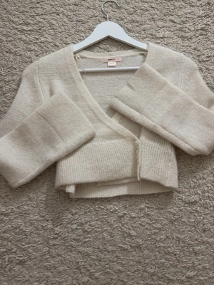 Krämvit cropped kofta Gina Tricot - Mysig krämvit cropped kofta från Gina Tricot med v-ringning och breda ärmar. ALDRIG ANVÄNT! Koftan har ribbstickade muddar och stängs med två knappar framtill. Perfekt att slänga över en topp för en chill look.