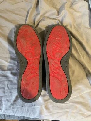 Gråa Louboutin sneakers med nitar - Säljer ett par gråa sneakers från Christian Louboutin med klassisk röd sula och coola silverfärgade nitar på hälen. Skorna är i mocka och har en låg profil med snörning. Perfekta för dig som vill sticka ut med exklusiv streetstyle.