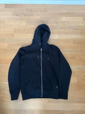 Svart zip-hoodie med fickor - En klassisk svart hoodie med dragkedja framtill, huva med dragsko och två stora fickor från Jack & Jones. Perfekt för en chill och avslappnad stil. Mjuk och skön bomullsblandning som passar året runt. Mycket fint skick. Storlek S. Pris kan alltid diskuteras✅.