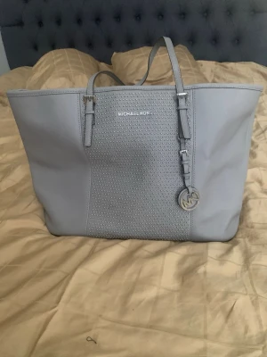 Ljusgrå handväska från Michael Kors - Snygg ljusgrå handväska från Michael Kors i stilren design. Väskan har silverfärgade detaljer, dekorativa nitar på framsidan och ett MK-hänge. Tillverkad i slätt konstläder med två axelband och gott om plats för dina grejer.