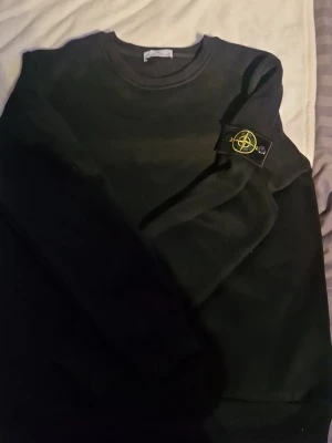 Svart sweatshirt från Stone Island - Säljer en svart sweatshirt från Stone Island med den klassiska logotypen på ärmen. Tröjan har rund halsringning och är tillverkad i mjukt bomullsmaterial. Perfekt för dig som gillar streetwear och vill ha en stilren look.