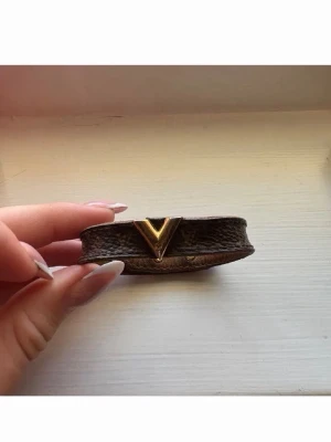 Louis Vuitton läderarmband - Snyggt brunt armband från Louis Vuitton i klassiskt monogramläder. Armbandet har en guldfärgad V-detalj framtill och tryckknappar i guld på baksidan. Perfekt accessoar för dig som gillar lyxiga detaljer och tidlös design. Nypris 3000
