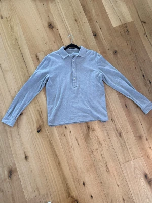 Grå långärmad henley från J.Lindeberg - Snygg grå långärmad pikétröja från J.Lindeberg med klassisk krage och knäppning. Tröjan är i mjukt material och har en stilren look som passar perfekt till jeans, chinos eller väst på varmare dagar. Passar M/S.