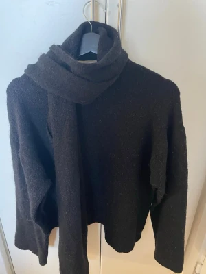 Svart stickad tröja med halsduk - Mysig svart stickad tröja med matchande halsduk. Tröjan har rund halsringning, långa ärmar och ribbade muddar. troligen ull eller en ullmix. Perfekt för kyliga dagar och enkel att styla.