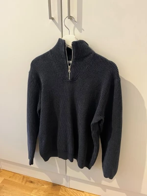 Mörkblå stickad tröja Cubus med dragkedja - Mörkblå stickad tröja i grov ribbstickning med hög krage och halvzip framtill. Tröjan har långa ärmar och en avslappnad passform, perfekt för lager på lager. Materialet ser ut att vara mjukt och värmande, troligen bomull eller ullblandning.