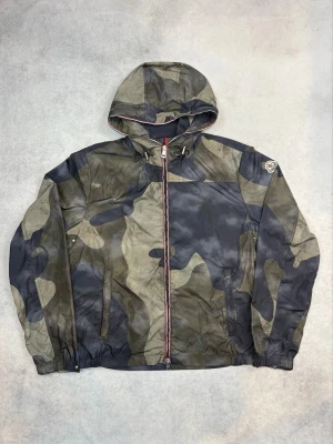 Moncler camo vindjacka med huva - Snygg vindjacka från Moncler i camo-mönster med dragkedja framtill och huva. Jackan har två sidofickor med dragkedja och elastiska muddar vid ärmslut och midja. Perfekt för dig som vill ha en streetig och trendig look.