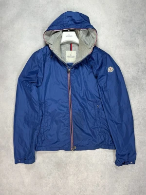 Blå vindjacka från Moncler - Snygg blå vindjacka från Moncler med huva och dragkedja framtill. Jackan har grått innerfoder, två sidofickor och Moncler-logga på ärmen. Detaljer i rött, vitt och blått längs dragkedjan ger en sportig vibe. Perfekt för blåsiga dagar.