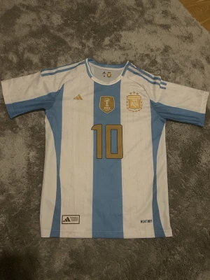 Argentina fotbollströja Messi #10 - Säljer en klassisk Argentina landslagströja från Adidas med Messi och nummer 10 i guld på ryggen. Tröjan är vit med ljusblå ränder, har korta ärmar och detaljer i guld. Tillverkad i lätt och ventilerande material, perfekt för fotboll eller för en soft dag. Aldrig använd!