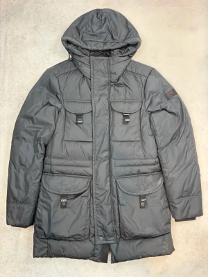 Mörkgrå parkas från Peuterey - Snygg mörkgrå parkas från Peuterey med fyra stora fickor framtill och huva. Jackan har långa ärmar och stängs med dragkedja och knappar. Perfekt för kalla dagar och har en klassisk, rak passform.