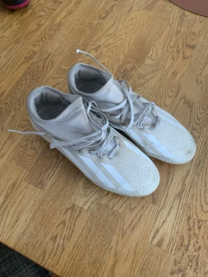 Vita Adidas fotbollsskor med dobbar - Snygga vita fotbollsskor från Adidas med klassiska tre ränder på sidorna. Skorna har högt skaft i strumpliknande material och snörning för bra passform. Utrustade med fasta dobbar för spel på gräs. Perfekta för dig som vill ha stilrena och funktionella skor på planen.