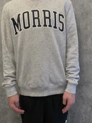 Grå sweatshirt från Morris - Klassisk grå sweatshirt från Morris med stor svart logotyp på bröstet. Tröjan har rund halsringning, ribbade muddar vid ärmslut och nederkant samt en avslappnad passform. Perfekt för en casual och sportig stil.