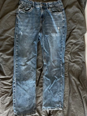 Blå raka jeans med slitningar - Säljer ett par klassiska blå jeans med raka ben och diskreta slitningar framtill. Jeansen har fem fickor, normal midja och är gjorda i denim. Perfekta för en avslappnad och trendig look.