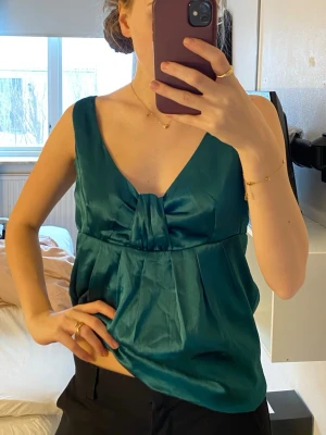 Grön sidenlinnetopp - festtopp - Snygg grön linnetopp i glansigt sidenmaterial med v-ringning och dekorativ rosett framtill. Toppen är ärmlös och har en lös passform som ger en avslappnad men chic look. Perfekt att styla med jeans eller kjol för en trendig outfit. I storlek S 