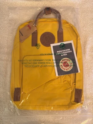 Gul Kånken ryggsäck från Fjällräven - Snygg gul Kånken ryggsäck från Fjällräven med klassisk rund logotyp i läder framtill. Ryggsäcken har bruna handtag och detaljer samt justerbara axelband. Tillverkad i slitstarkt material, perfekt för skolan eller utflykter.