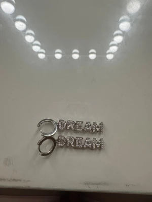 Silvriga örhängen med text DREAM - Säljer ett par silvriga örhängen i hoops-modell med ordet DREAM i stora bokstäver. Bokstäverna är täckta av små glittrande stenar som ger extra bling. Perfekta för dig som vill sticka ut och visa din personlighet med accessoarer.
