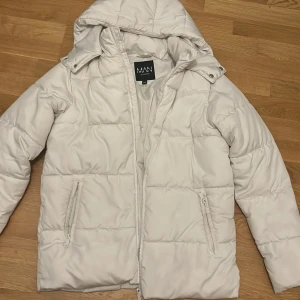Vit pufferjacka - Snygg vit pufferjacka från MAN med dragkedja framtill och två sidofickor med dragkedja. Jackan har en vadderad design och en stor huva med tryckknappar. Perfekt för kalla vinterdagar och riktigt skön att bära.