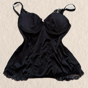 Svart korsettlinne med spetsdetaljer - Snyggt svart korsettlinne med tunna axelband och spetsdetaljer vid byst och nederkant. Dekorativ liten rosett framtill och figurnära passform som framhäver siluetten. Perfekt att styla med både jeans och kjol.
