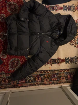 Svart pufferjacka från Polo Ralph Lauren - Säljer en svart pufferjacka från Polo Ralph Lauren med huva och dragkedja. Jackan har det klassiska röda Polo-logot på bröstet och stora quiltade sektioner för extra värme. Perfekt för kalla vinterdagar och riktigt snygg streetstil.