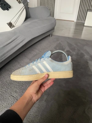 Ljusblå Adidas sneakers i mocka - Snygga ljusblå sneakers från Adidas med klassiska vita ränder på sidorna. Skorna är i mjuk mocka och har en beige sula som ger en retrovibe. Perfekta för dig som gillar stilrena och bekväma sneakers med ikonisk design.