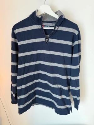 Randig blå tröja från Chaps - Mörkblå långärmad tröja från Chaps med gråa horisontella ränder. Tröjan har halv dragkedja framtill och ståkrage. Tillverkad i mjukt bomullsmaterial, perfekt för en avslappnad stil.