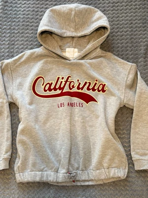 Grå hoodie med California-tryck från Zara - Snygg grå hoodie med huva och trycket 'California Los Angeles' i rött och gult framtill från Zara. Mjuk och skön bomullsblandning, ribbade muddar och dragsko nedtill för extra passform. Perfekt för en avslappnad streetwear-look.
