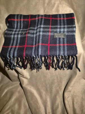 Rutig halsduk från Burberry - Mörkblå halsduk från Burberry i klassiskt rutigt mönster med röda och grå ränder. Halsduken har tvinnade fransar i ändarna och en tygpatch med Burberry-loggan. Perfekt accessoar för att lyfta din outfit med en tidlös brittisk stil.