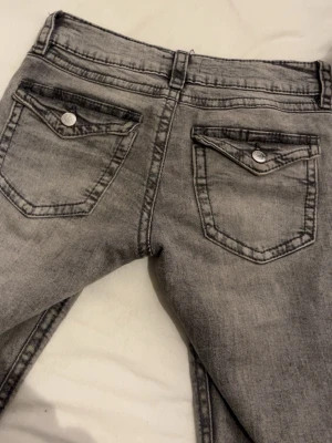 Grå slim fit jeans med ficklock - Snygga grå slim fit jeans med slitningseffekt och ficklock bak. Klassisk femficksmodell med silverfärgade knappar och bälteshällor. Jeansen har en modern tvättad look och smal passform som passar perfekt till en trendig vardagsstil.