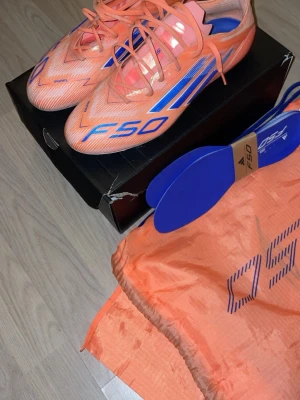 Adidas F50 orange/blå fotbollsskor - Säljer ett par Adidas F50 fotbollsskor i en riktigt snygg orange färg med blå detaljer och snörning. Skorna har en låg profil och är tillverkade i lätt syntetmaterial för maxad speed på planen. Medföljer matchande skopåse och extra innersulor, endast använda 2 träningar.