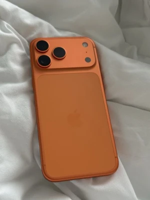Iphone 17 pro max 512GB - Snygg orange iPhone med stilren design och tredubbel kamera på baksidan. Telefonen har rundade hörn och är tillverkad i glas och aluminium. Perfekt för dig som vill sticka ut med en unik färg och ta grymma bilder.
