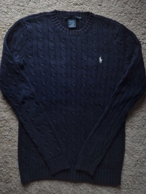 Mörkblå kabelstickad tröja Polo Ralph Lauren - Kabelstickad mörkblå tröja från Polo Ralph Lauren med rund halsringning och den klassiska broderade logotypen i vitt på bröstet. Tröjan har långa ärmar och ribbade muddar vid ärmslut och nederkant. Perfekt för en stilren och avslappnad look.