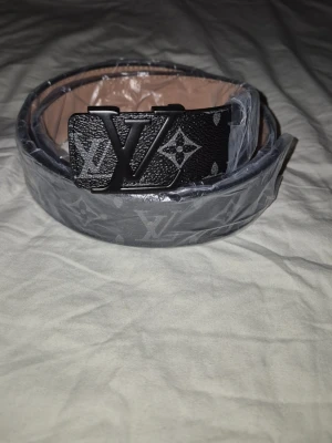 svart monogram skärp - Snyggt svart skärp med klassiskt monogrammönster silver på spännet. Bältet har en stilren design och passar perfekt till jeans eller kostymbyxor. Kommer med originalförpackning och kvitto