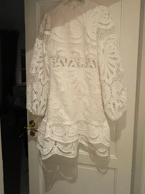 Vit spetsklänning med volang - Säljer en vit kortklänning i spets med långärmade ärmar och volangdetaljer. Klänningen har ett vackert broderat mönster och hög hals. Perfekt för studenten!