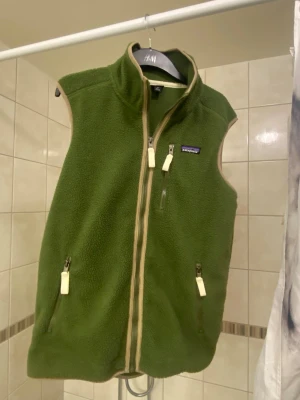 Grön fleeceväst från Patagonia - Säljer en grön fleeceväst från Patagonia med beige detaljer och dragkedja framtill. Västen har två fickor med dragkedja och ett klassiskt Patagonia-märke på bröstet. Perfekt för lager-på-lager och riktigt skön att bära. Hör gärna av dig