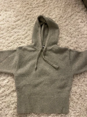 Grå stickad hoodie med huva - Mysig grå stickad hoodie från NA-KD med ribbad kant nedtill och dragsko i huvan. Perfekt för chill dagar och enkel att matcha med jeans eller mjukisbyxor. Materialet känns mjukt och värmande, och modellen är lite croppad för en trendig look.