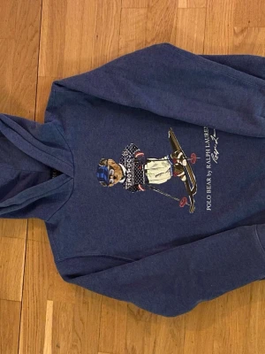 Blå hoodie Polo Ralph Lauren björn - Mörkblå hoodie från Polo Ralph Lauren med ikoniskt Polo Bear-tryck på bröstet där björnen har skidutrustning. Mjuk bomullsblandning och klassisk huva. Perfekt för dig som gillar streetwear och vill ha en unik detalj i garderoben.