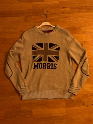 Beige stickad tröja Morris - Stickad beige tröja från Morris med Union Jack-flagga och texten 'Morris' på bröstet. Tröjan har ribbstickade muddar, rund hals och dekorativa förstärkningar på armbågarna. Perfekt för dig som gillar klassisk och preppy stil.