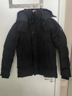 Canada Goose Wyndham black label - Säljer en svart dunjacka från Canada Goose med stor huva och hög krage. Jackan har två stora dragkedjeförsedda fickor framtill, ribbade muddar vid ärmsluten och en patch på ärmen. Perfekt för kalla vinterdagar.
