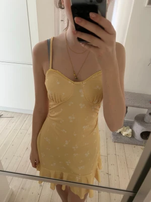 Gul blommig kortklänning med volang - Söt gul kortklänning med små vita blommor och volangkant nertill. Klänningen har smala axelband och markerad byst med söt spetsdetalj vid urringningen. Perfekt för varma dagar och ger en fräsch sommarkänsla. Köpte på plick och vet ej märket