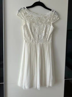 Vit spetsklänning från Vila - Superfin vit klänning med spetsdetaljer och transparent övre del. Klänningen har korta ärmar, blommigt spetsmönster och en lätt, flödande kjol. Perfekt för sommarens alla tillfällen och ger en romantisk vibe. Använd en gång!