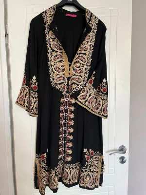 Svart broderad klänning/tunika Indiska - Elegant svart klänning/tunika från Indiska med vackra broderier i guld, beige och vinröd. Klänningen har vida ärmar och detaljerad snörning vid halsen. Perfekt för dig som gillar unika och färgstarka plagg med etnisk touch.