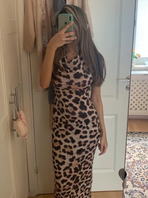 Leopardmönstrad långklänning - Säljer en snygg långklänning med leopardmönster. Klänningen har en mjuk, åtsittande passform och en draperad halsringning. Perfekt för dig som vill sticka ut med ett djuriskt print. Det är även öppenrygg. Materialet känns stretchigt och bekvämt.