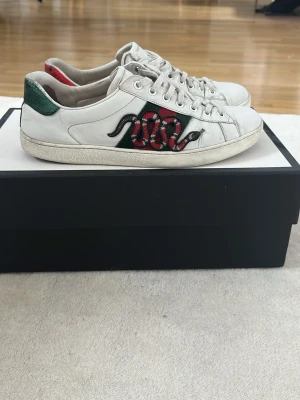 Gucci Ace sneakers med orm - Säljer ett par vita Gucci Ace sneakers med ikonisk broderad orm på sidan. Skorna är tillverkade i läder och har klassisk låg profil med snörning. Perfekt för dig som vill sticka ut med en lyxig stil.