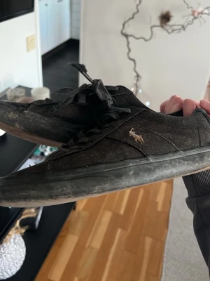 Svarta Polo Ralph Lauren sneakers - Svarta sneakers från Polo Ralph Lauren med klassisk låg profil och broderad logga på sidan. Skorna har svarta snören, gummisula och är tillverkade i canvasmaterial. Perfekta för en avslappnad och stilren look.