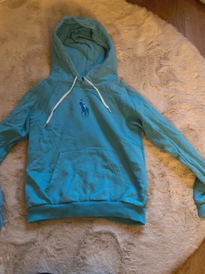 Turkos hoodie från Polo Ralph Lauren - Turkos hoodie från Polo Ralph Lauren med klassisk logga på bröstet. Hoodien har vit dragsko, stor huva och en praktisk magficka. Materialet är mjukt och bekvämt, perfekt för chill dagar. Snygg och enkel att matcha med jeans eller joggers.