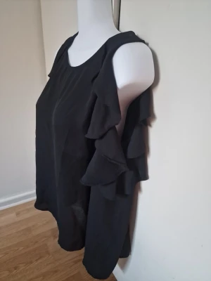 Svart blus med volangärmar - Svart blus med "cold shoulders" och volangärmar. Materialet är lätt och luftigt, perfekt för varma dagar.