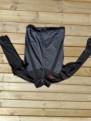 Svart träningsjacka från Puma - Säljer en svart träningsjacka från Puma med hel dragkedja och diskret grå panel på framsidan. Jackan har långa ärmar och ett rött Puma-logo på bröstet. Perfekt för löpning eller gym. Materialet är syntetiskt och lätt, vilket gör den smidig att bära.