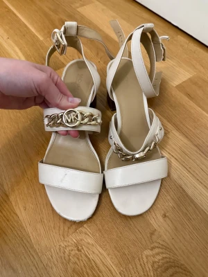 Vita sandaler med klack från Michael Kors - Snygga vita sandaler från Michael Kors med bred rem framtill, guldfärgad kedjedetalj och MK-logga. Klack i trälook och justerbara remmar runt vristen. Perfekta för att lyfta din outfit med en lyxig touch.