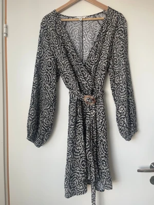 omlottklänning med bälte - Snygg svartvit omlottklänning med lövliknande mönster och markerad midja med bälte i plastspänne. Klänningen har lång ärm och v-ringad hals. Perfekt för dig som gillar trendiga och bekväma plagg med fina detaljer.